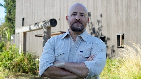 man standing with arms cross with longhouse in background